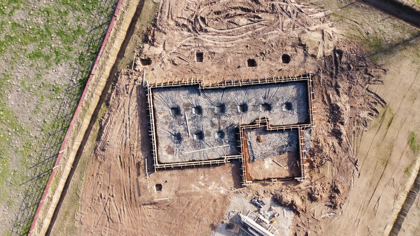 Top-down view of a foundation under construction: excavated ground, timber edge formwork, a polythene membrane and pile cap pits.