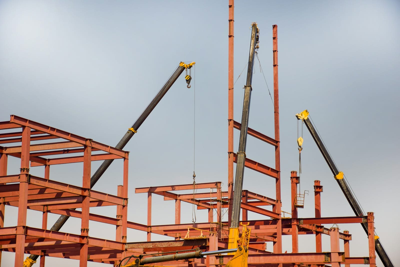 A multi-storey structural steel frame under erection, with bolted connections and three mobile crane booms against an overcast sky.