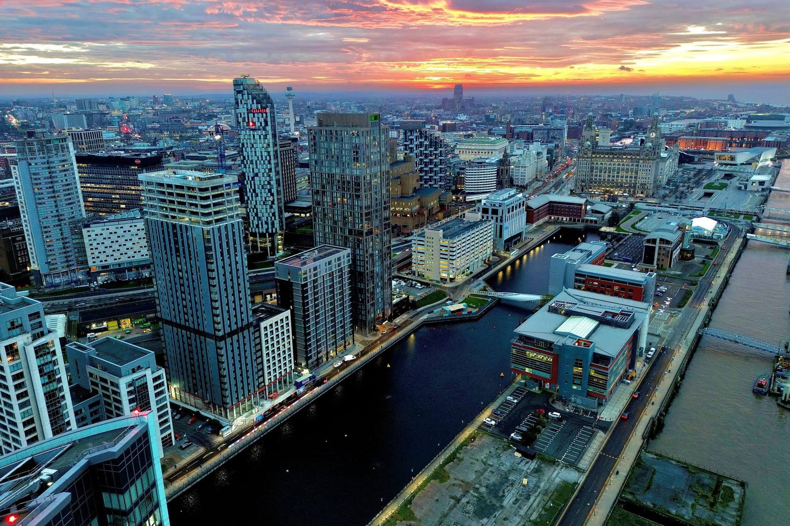 A city waterfront at dusk from the air, towers, docks and a river under a red sky.